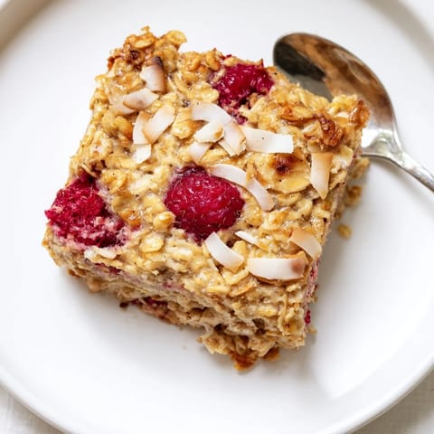 Duftendes Baked Oatmeal mit saftigen Himbeeren und knusprigem Kokos, serviert warm mit Ahornsirup für ein herzhaftes Frühstück.